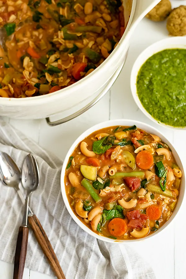 Overhead shot of a bowl of vegan minestrone soup with the pot of soup and pesto in the background. Two wooden handled spoons resting on a grey striped napkin to the left of the bowl. 