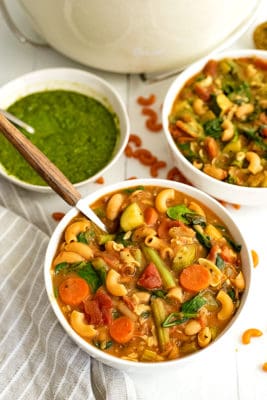 Large white bowl filled with vegan minestrone pesto with a wooden handled spoon in the bowl. A bowl of pesto and a second bowl of soup in the background