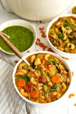 Large white bowl filled with vegan minestrone pesto with a wooden handled spoon in the bowl. A bowl of pesto and a second bowl of soup in the background
