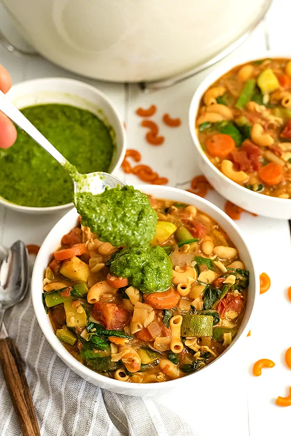 Spoonful of pesto being added to a bowl of vegan minestrone soup with the bowl of pesto in the background