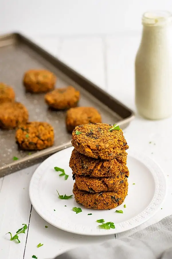 Sweet potato patties stacked on a white plate with a sheet pan full of sweet potato patties and a bottle of ranch dressing in the background