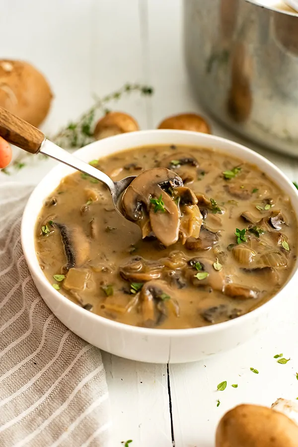 Large white bowl filled with the best ever mushroom soup. A wooden handled spoon is holding up a spoonful of soup. There is a grey striped napkin to the left of the bowl and the silver pot in the background