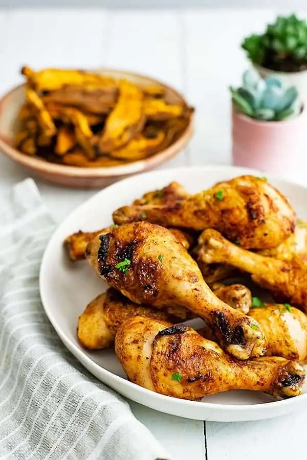 Close up of a large white plate filled with baked chicken drumsticks. Plate of baked sweet potato fries in the background