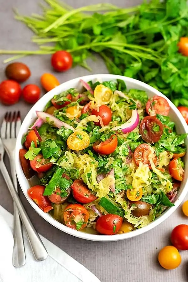 Overhead shot of a large white bowl filled with Greek Spaghetti Squash Salad. Tomatoes and parsley surround the bowl and there is a white napkin and two forks off to the left.