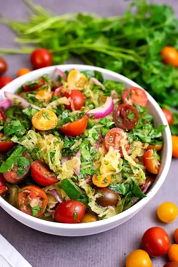 Close up of a large white bowl filled with Greek Spaghetti Squash Salad. There are tomatoes around the bowl and parsley leaves in the background.