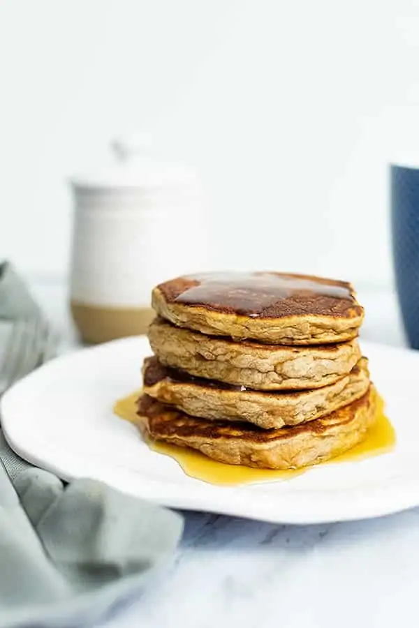 Stack of gluten free protein pancakes with syrup dripping down. White ceramic jar in the background