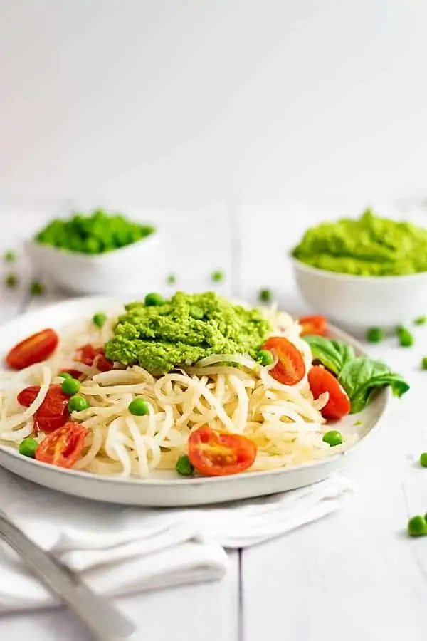 Plate filled with pasta noodles and tomatoes topped with pea pesto. Bowl of pea pesto in the background
