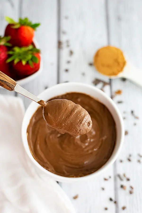 Overhead shot of a spoonful of the chocolate protein pudding over the bowl of pudding. Strawberries and cacao powder in the background