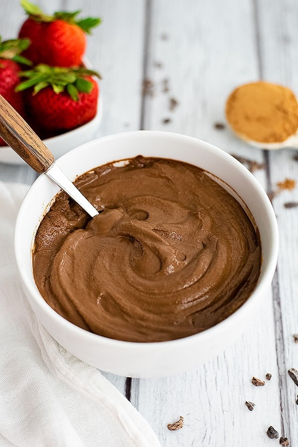Overhead shot of a bowl of chocolate protein pudding with a wooden handled spoon in the bowl. Strawberries in the background