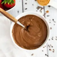 Overhead shot of a bowl of chocolate protein pudding with a wooden handled spoon in the bowl. Strawberries in the background