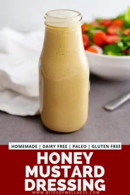 Glass bottle filled with homemade honey mustard dressing recipe with a salad in the background. Pinterest text on the bottom