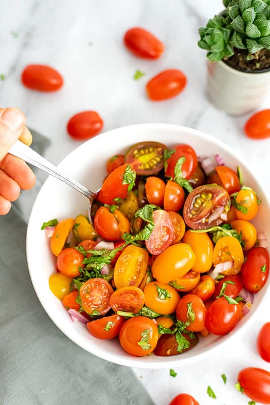 Bowl filled with tomato basil salad with a silver spoon being held into the bowl Cherry tomatoes surround the bowl.