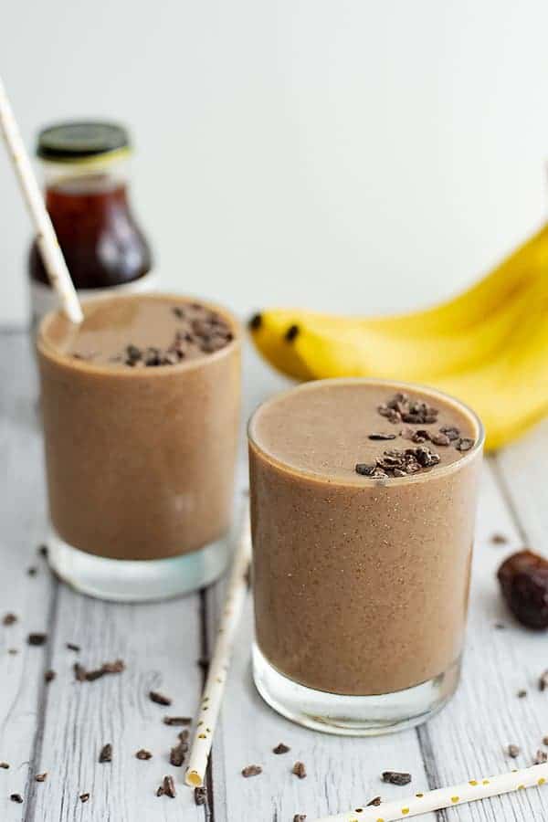 Two glasses of coffee banana protein smoothies with bananas and cold brew coffee in the background