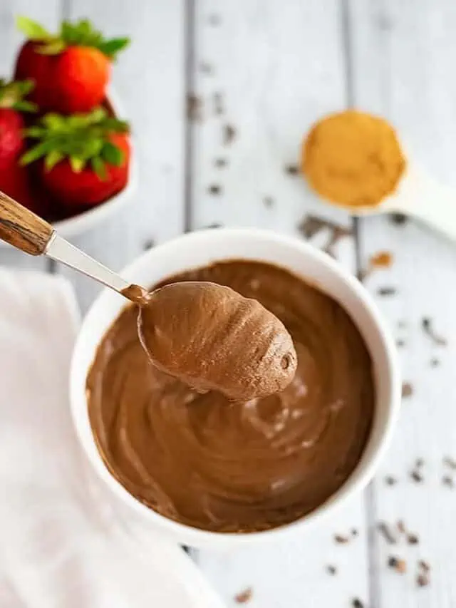 Overhead shot of a spoonful of the chocolate protein pudding over the bowl of pudding. Strawberries and cacao powder in the background