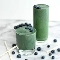 Two glasses of spinach blueberry smoothie with blueberries surrounding them