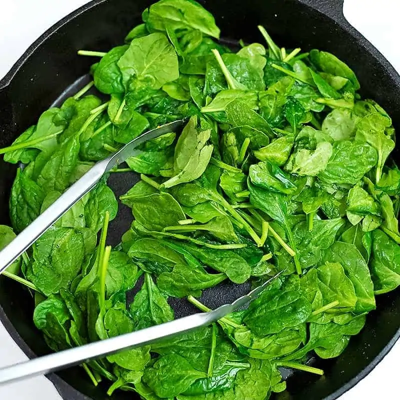 Tongs tossing arugula in cast iron skillet.