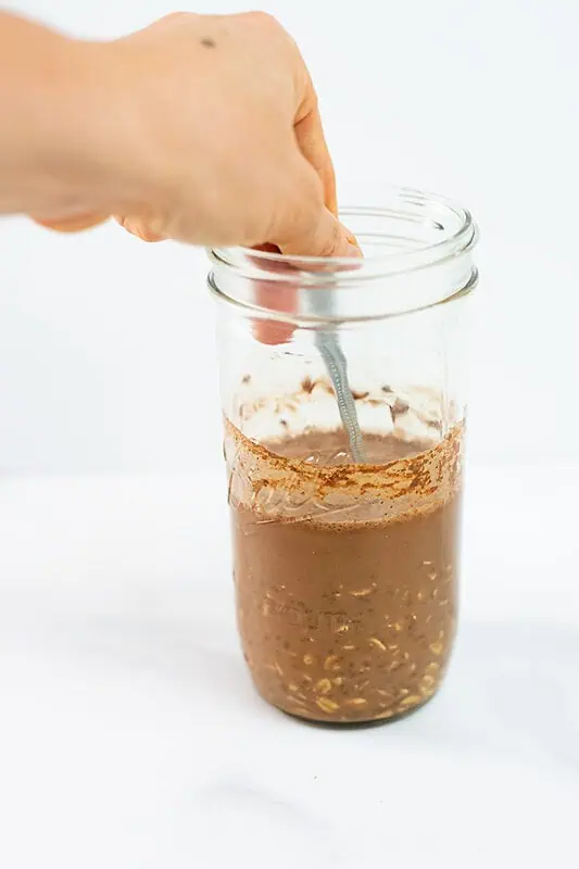 Hand holding spoon stirring protein powder into jar of oats.