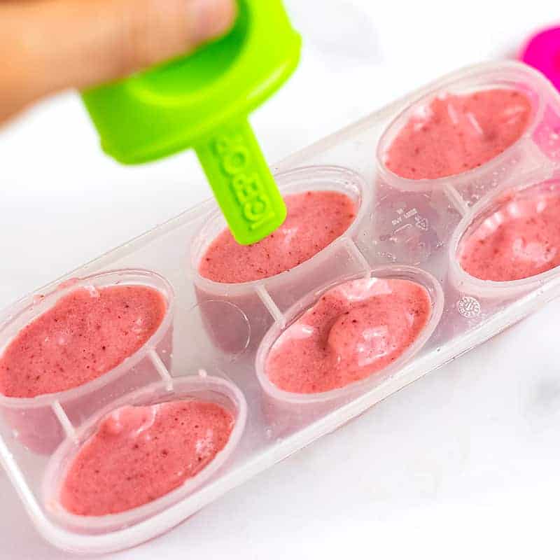 Hand putting the popsicle handle in the strawberry yogurt mixture
