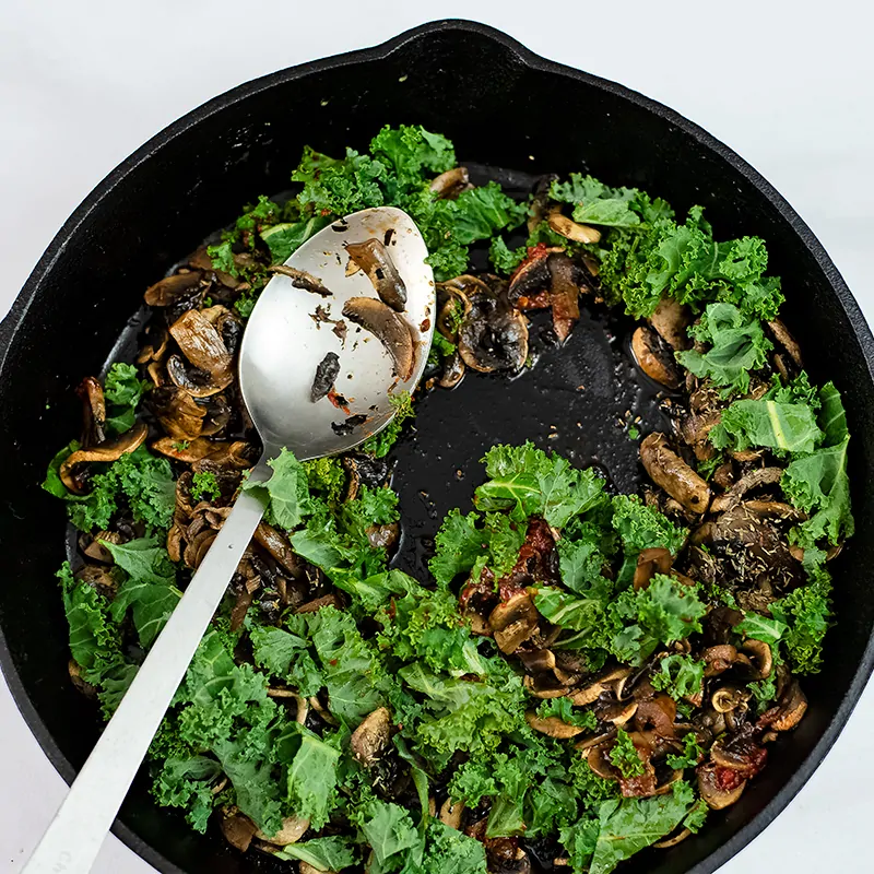 Fresh arugula wilting on cooked mushrooms in a skillet. 