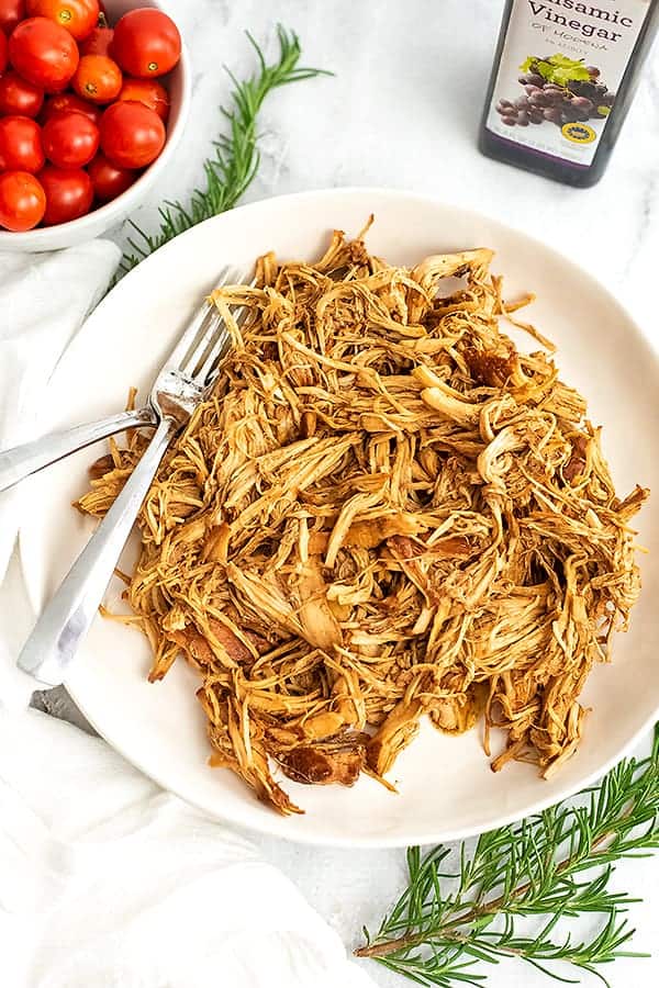 White plate filled with shredded balsamic chicken, tomatoes in background.