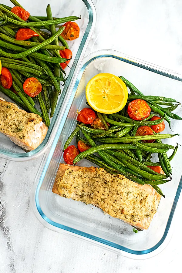 Glass container filled with tahini crusted salmon, green beans, tomatoes and a lemon.