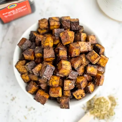 Bowl of baked balsamic tofu with a spoon of Italian seasoning below.