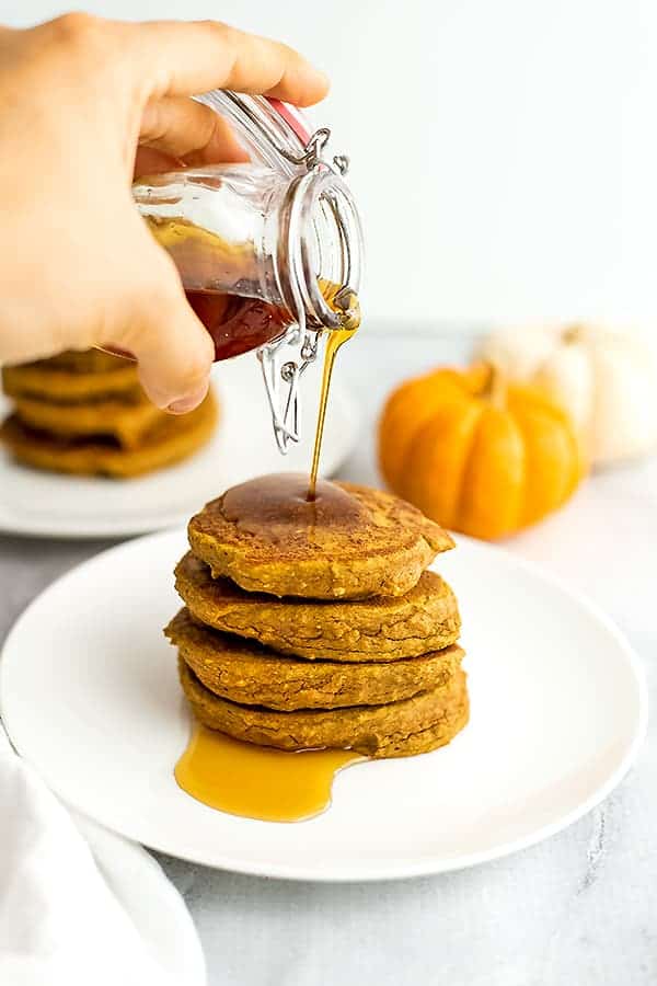 Syrup being poured over a stack of pumpkin pancakes on a white plate.