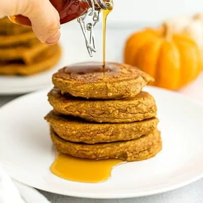 Syrup being poured over a stack of pumpkin pancakes on a white plate.