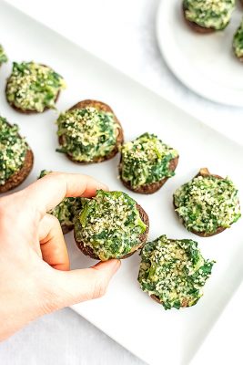 Hand picking up a spinach artichoke stuffed mushrooms from white plate.