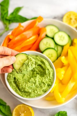 Cucumber being dipped into a bowl filled with pesto hummus.