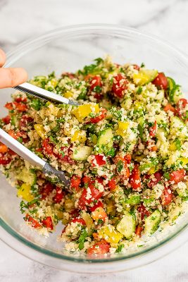 Tongs tossing the cauliflower tabbouleh in glass bowl.