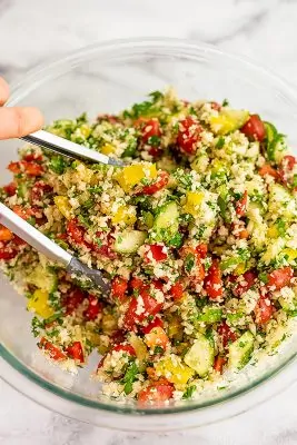 Tongs tossing the cauliflower tabbouleh in glass bowl.