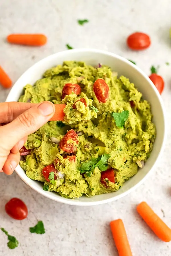 Baby carrot being dipped in large bowl of guacamole.