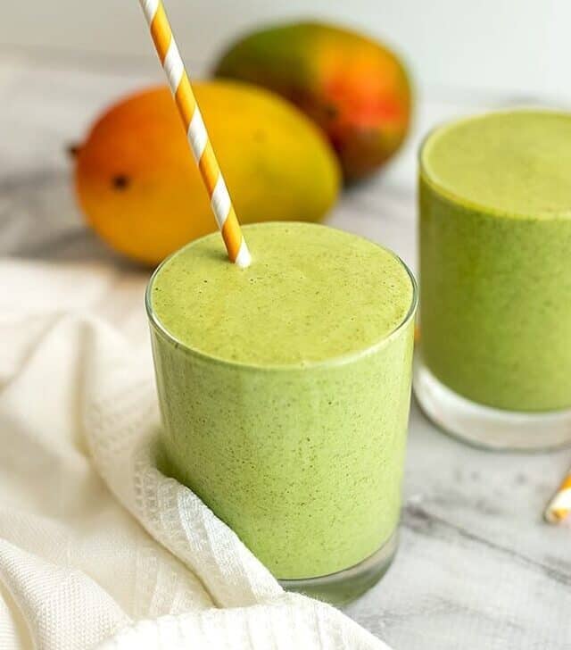 Two glasses of mango kale smoothie with mango in background.