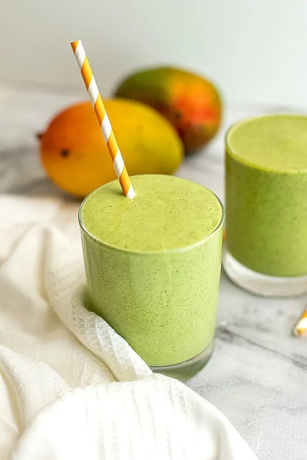 Two glasses of mango kale smoothie with mango in background.