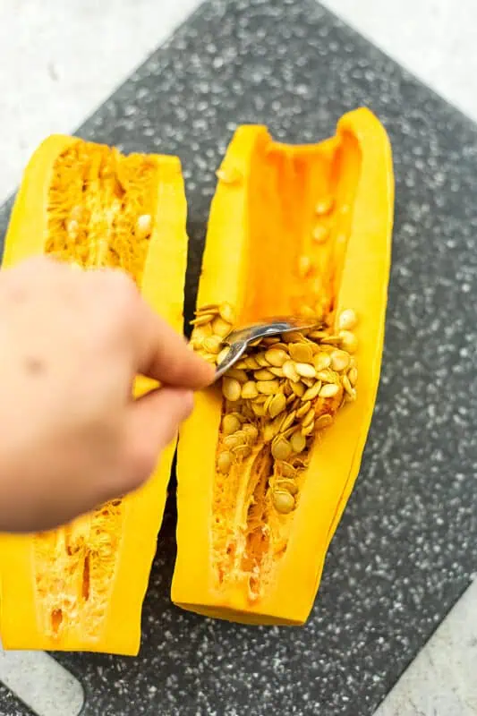 Hand holding a spoon scooping out the seeds from a delicata squash half.
