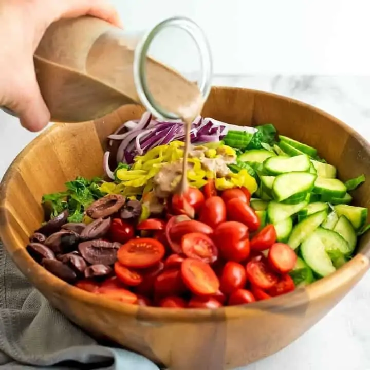 Greek salad in a wooden bowl with dressing being poured over the salad.