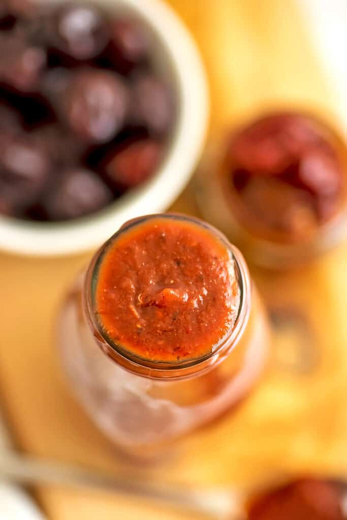 Glass bottle filled with chipotle cherry bbq sauce with cherries in background.