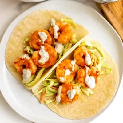Two buffalo grilled shrimp tacos with ranch drizzle on a plate.