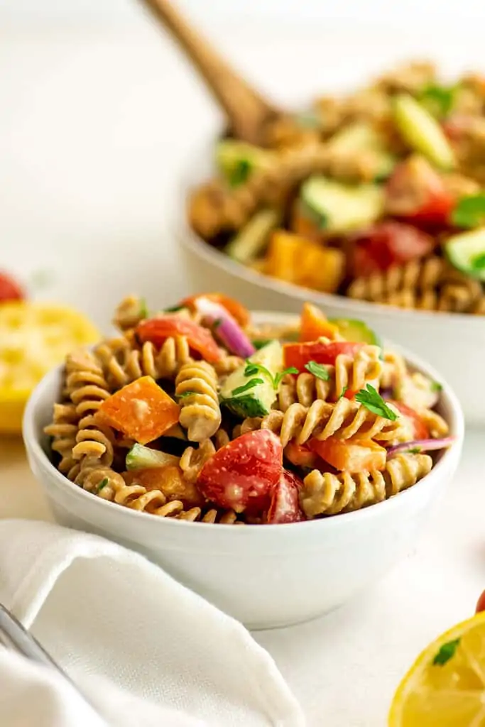 Hummus pasta salad in small white bowl with larger bowl in background.