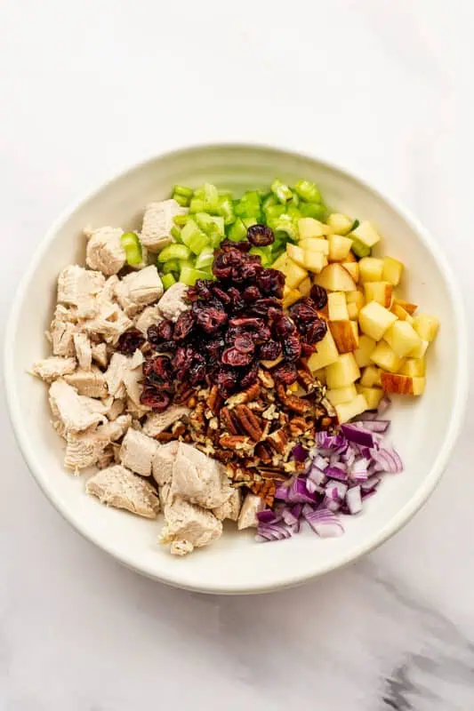 White bowl filled with chopped chicken, pecans, apples, celery red onion and cranberries.