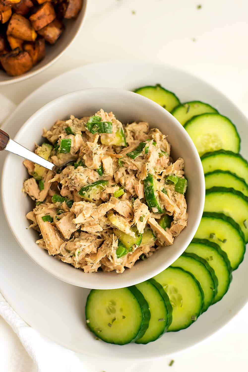 Tuna salad in a bowl with spoon in bowl and cucumbers on plate. 