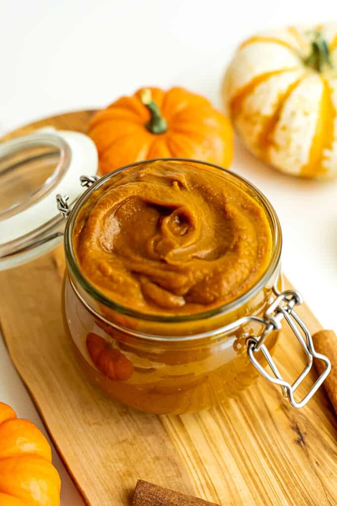 Pumpkin butter in glass jar over wooden plate, pumpkins in background.