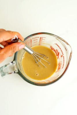Hand holding whisk in cup of maple tahini dressing.