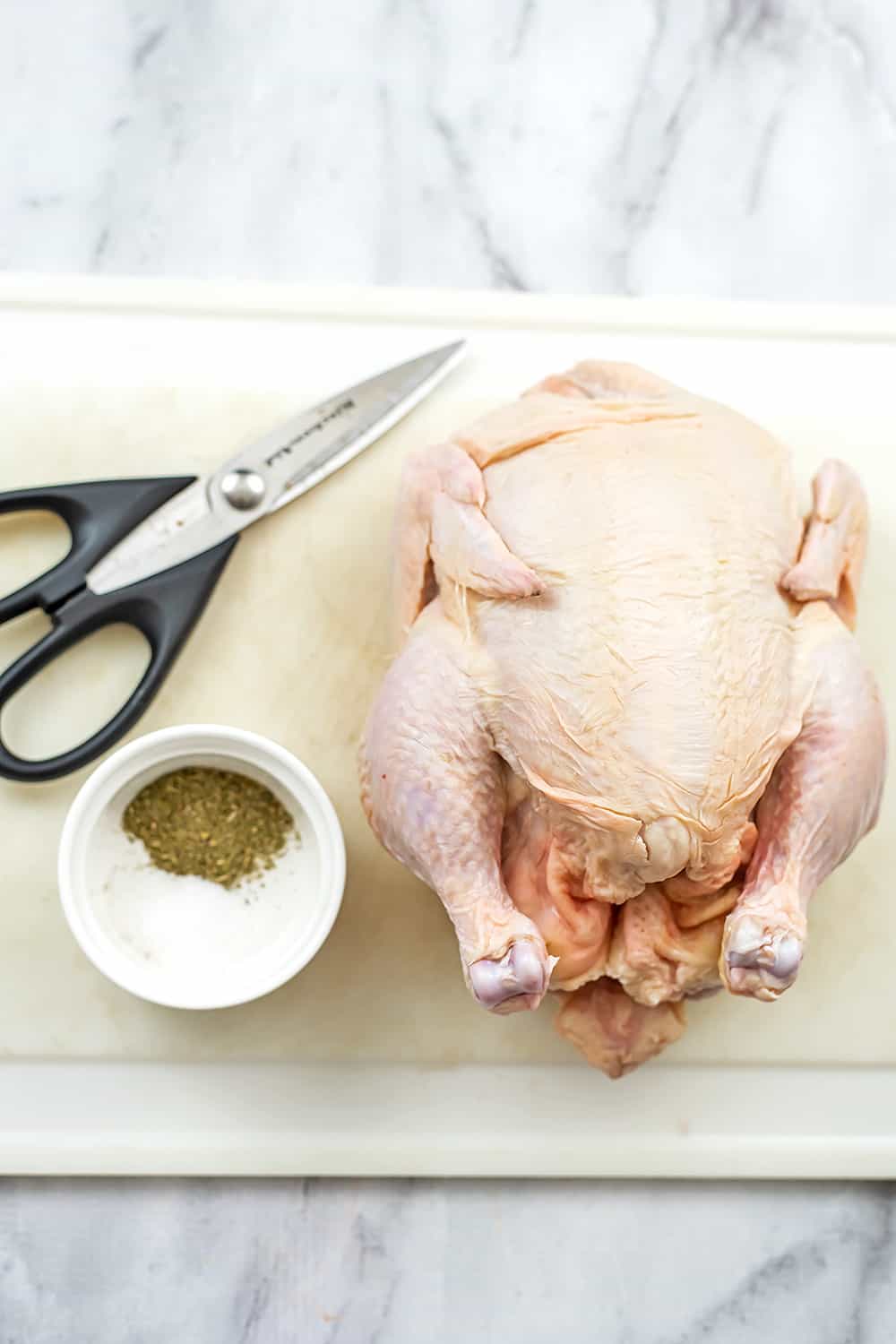 Whole chicken, kitchen shears and spices on white cutting board.