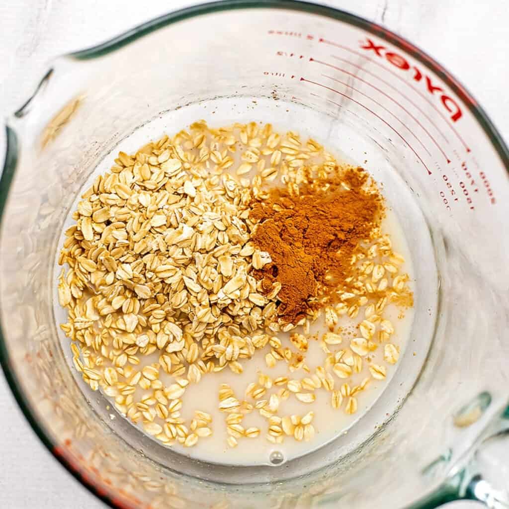Oats and cinnamon added to dairy free milk mixture in glass mixing bowl.