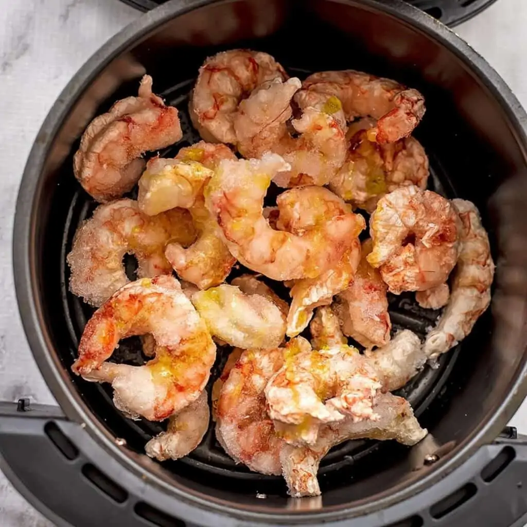 Frozen shrimp with olive oil drizzled on top in air fryer basket.