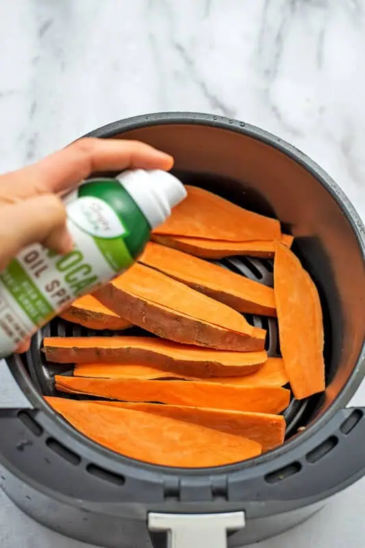 Olive oil being sprayed on the sweet potato wedges in air fryer basket.