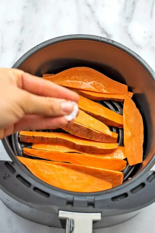 Salt being sprinkled on sweet potato wedges in air fryer basket.