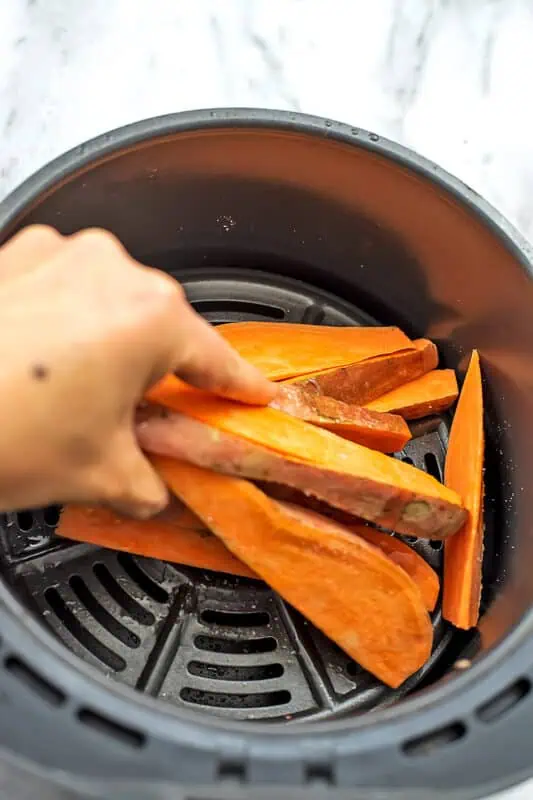 Hand moving around sweet potato wedges in air fryer basket.